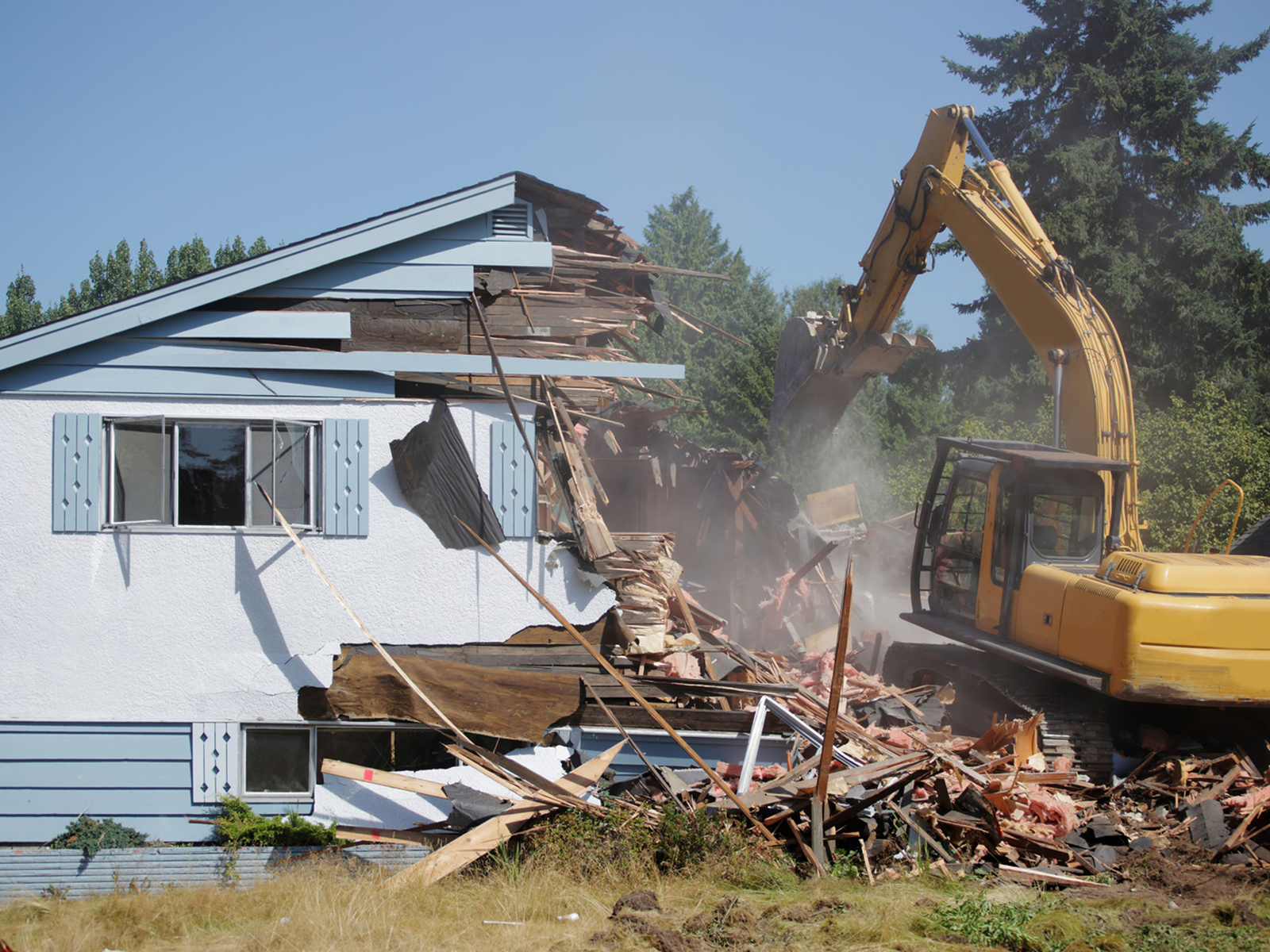 Excavator demolishing a blue wooden house.